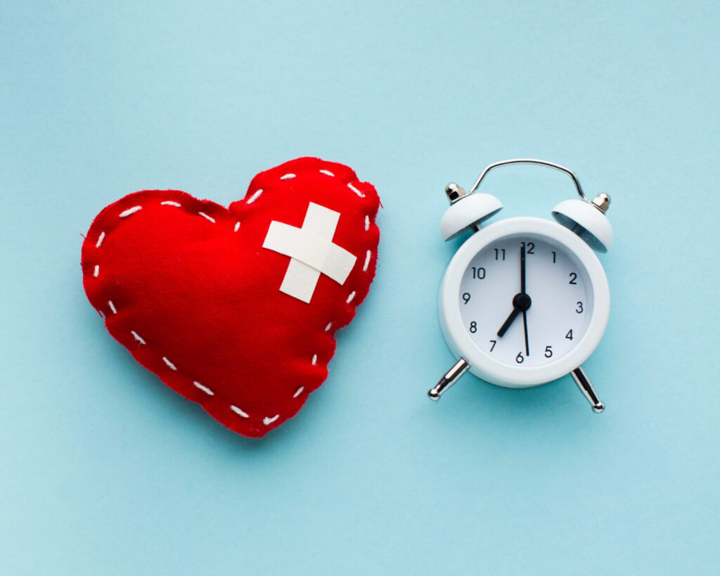 un cuore rosso con due cerotti a forma di x che coprono una ferita e una sveglia bianca poggiate su uno sfondo azzurro