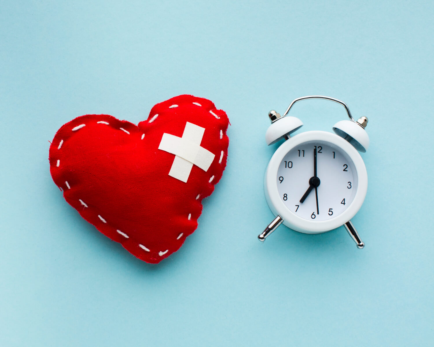 un cuore rosso con due cerotti a forma di x che coprono una ferita e una sveglia bianca poggiate su uno sfondo azzurro