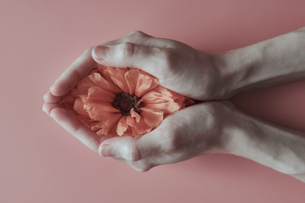 ue mani che avvolgono delicatamente un fiore arancione su sfondo rosa, simbolo di cura e femminilità, in riferimento alla Giornata Mondiale della Menopausa